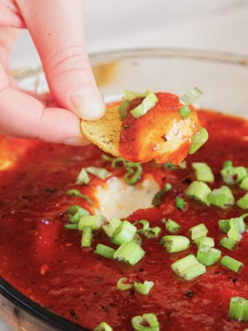 A hand dips a chip into a bowl of cheesy dip topped with salsa and chopped green onions.