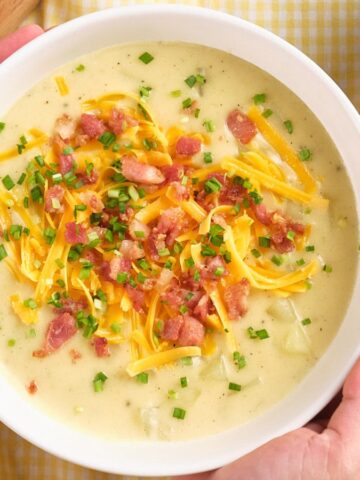 Bowl of creamy potato soup topped with cheddar cheese, bacon bits, and chopped chives, held in two hands.