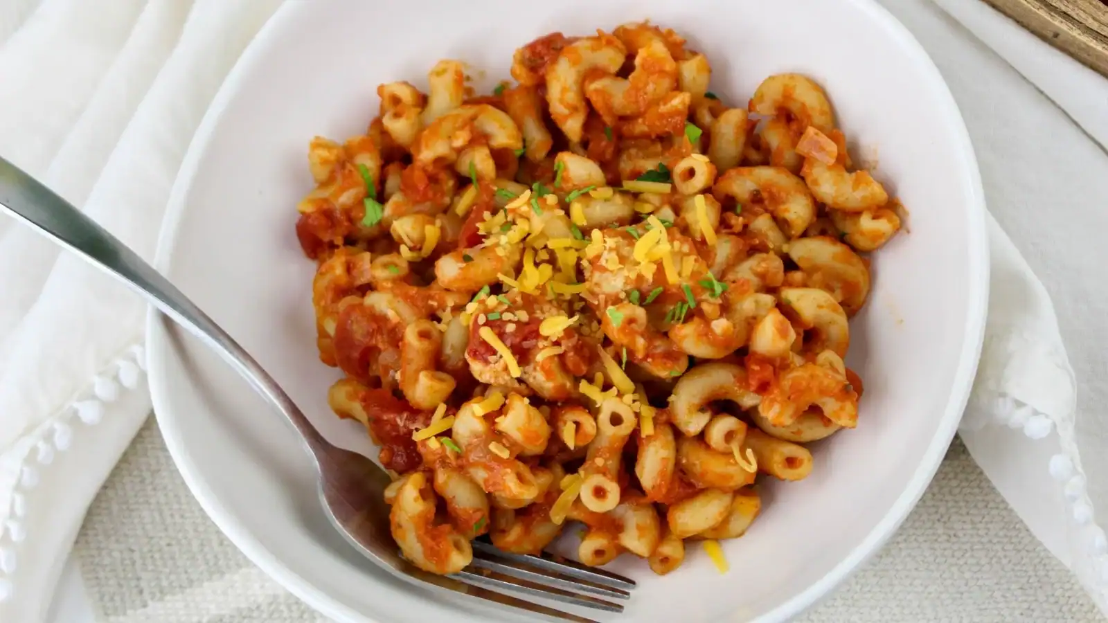 A white bowl of macaroni pasta in tomato sauce, topped with grated cheese and herbs, placed on a white cloth. A fork rests on the side of the bowl.