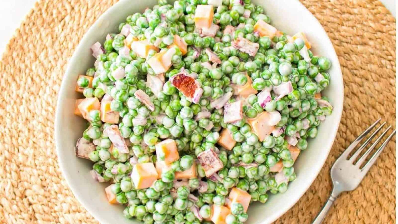 A bowl of pea salad with diced cheese, bacon pieces, and creamy dressing on a woven placemat, next to a fork.