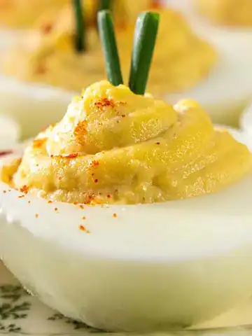 Close-up of deviled eggs topped with green herb garnish and a sprinkle of paprika, arranged on a plate with a floral pattern. The eggs are halved with yellow filling piped in the center.