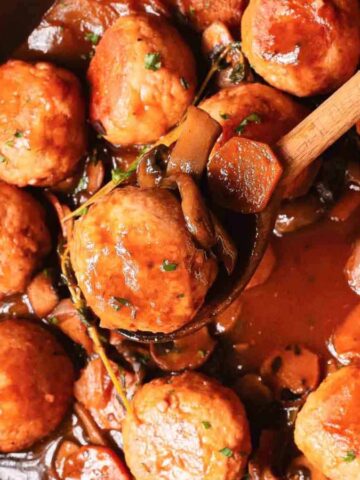 Round meatballs in a rich brown sauce with vegetables, served in a pan with a wooden spoon.