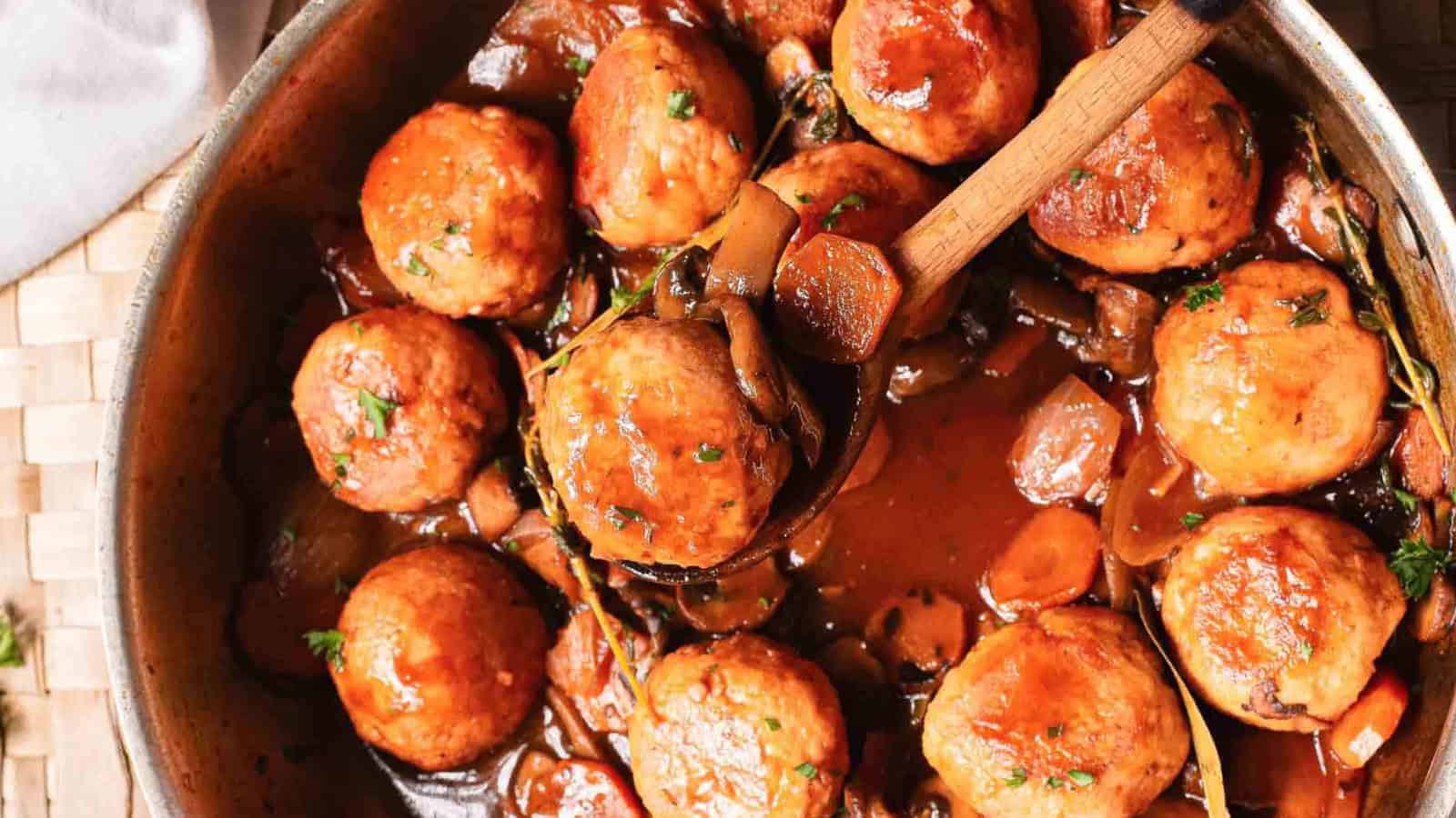 Round meatballs in a rich brown sauce with vegetables, served in a pan with a wooden spoon.
