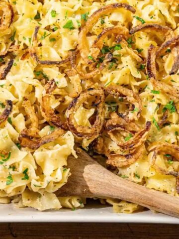 A baking dish filled with creamy noodle casserole, topped with crispy fried onions and chopped herbs, with a wooden spoon resting inside the dish.