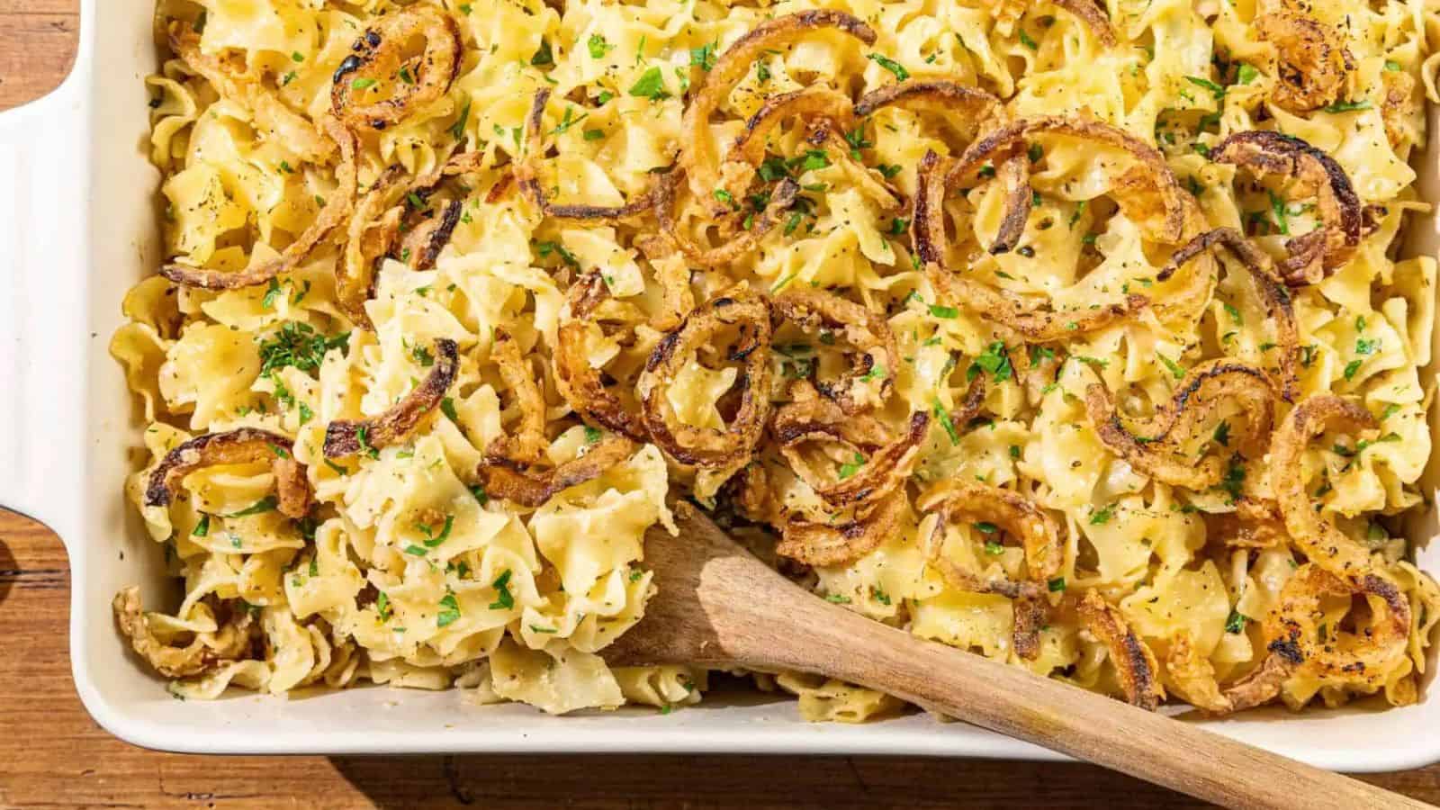 A French Onion Noodle Casserole filled with creamy egg noodles, topped with crispy fried onions and chopped parsley, sits invitingly in a dish with a wooden spoon resting inside.