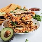 A stack of cheesy quesadillas with black beans and corn, garnished with cilantro, next to avocado and salsa.
