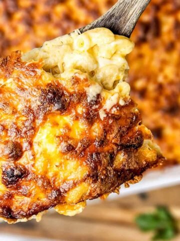 A wooden spoon holds a cheesy, golden-brown serving of baked macaroni and cheese above a casserole dish.