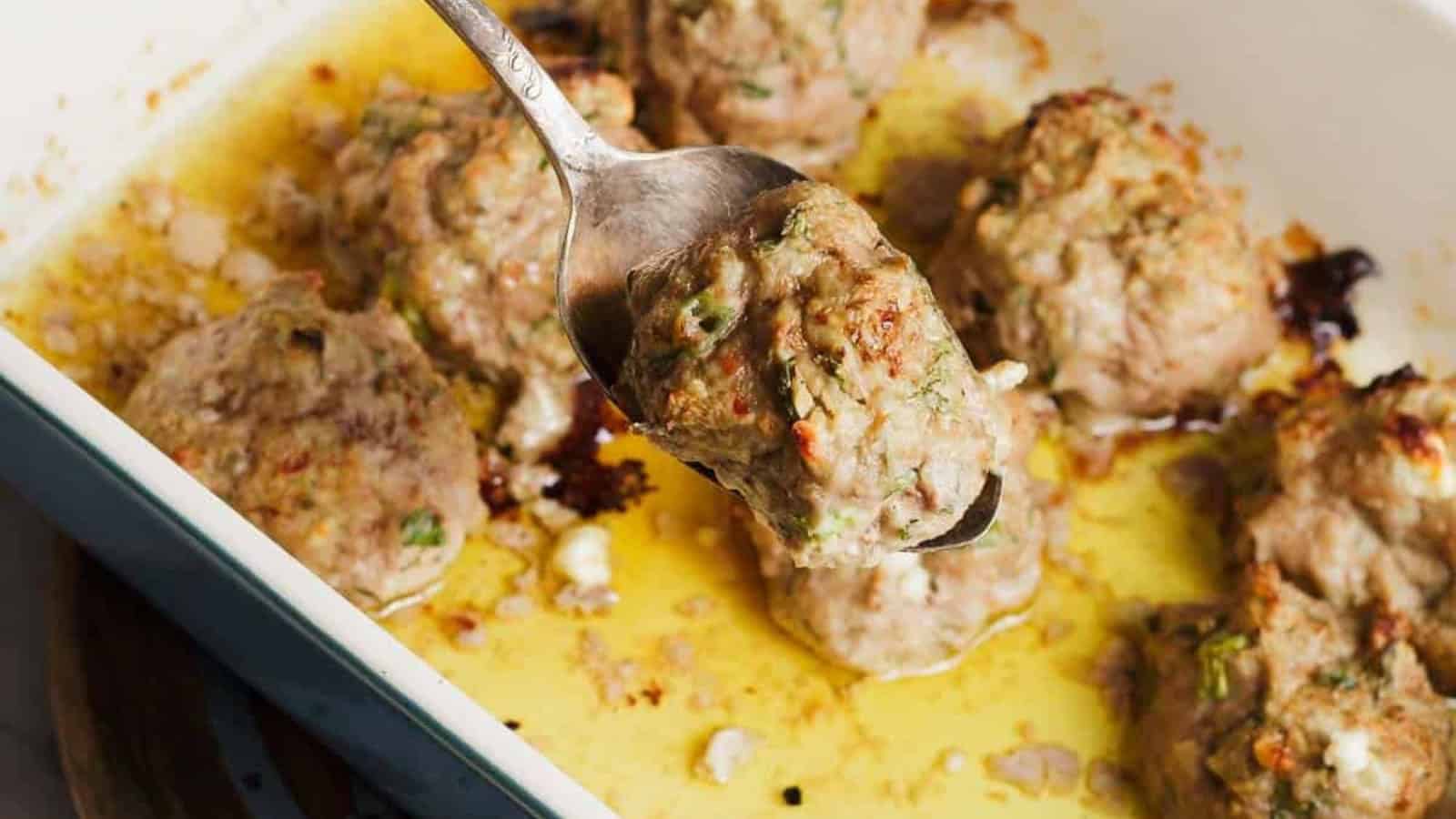 A spoon lifts a baked meatball from a dish filled with several meatballs in a golden, oily sauce.