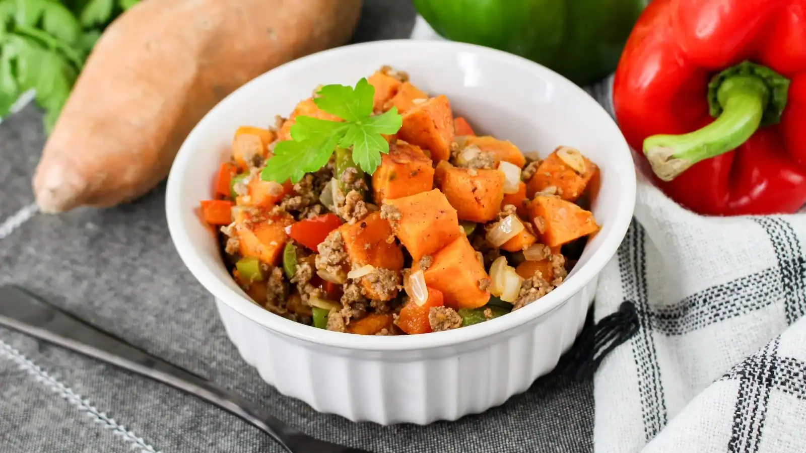 A white bowl filled with a sweet potato and ground beef dish, garnished with a green herb. Surrounding the bowl are a sweet potato, green pepper, and red pepper on a cloth.