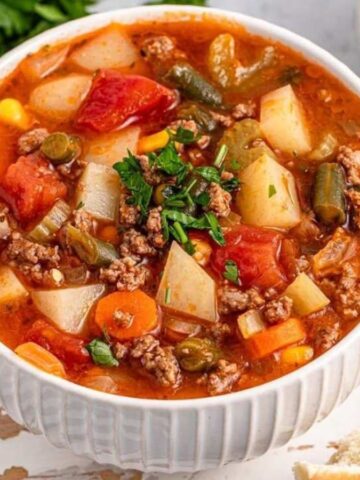 A bowl of hearty vegetable beef soup with bread rolls, garnished with fresh herbs, on a white table.