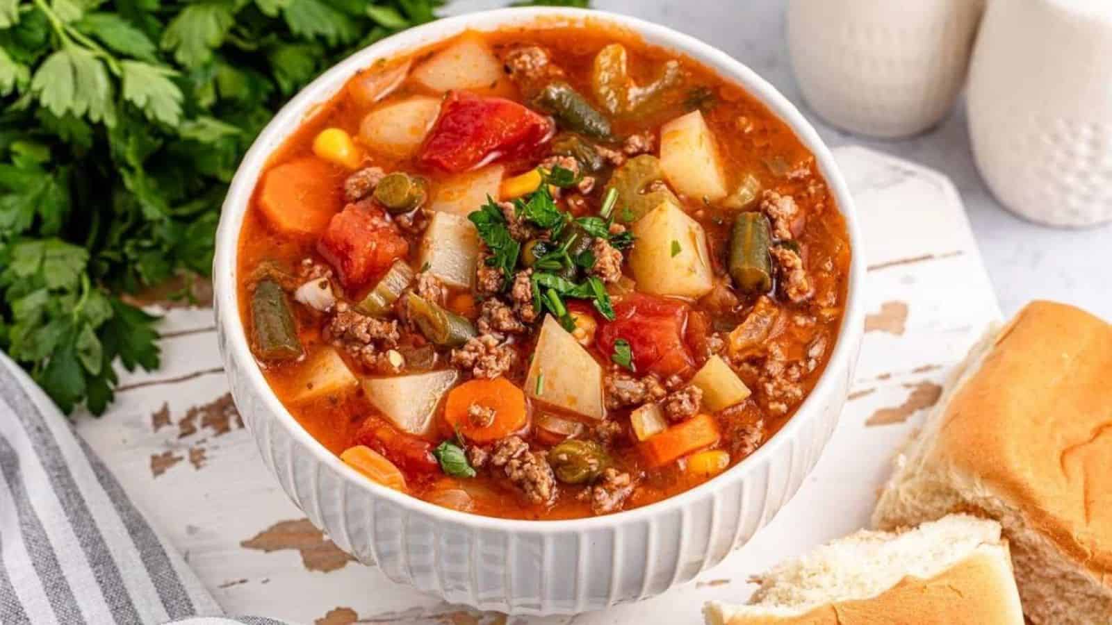 A bowl of vegetable beef soup with potatoes, carrots, green beans, corn, and ground beef, garnished with parsley. A dinner roll and parsley are beside the bowl.