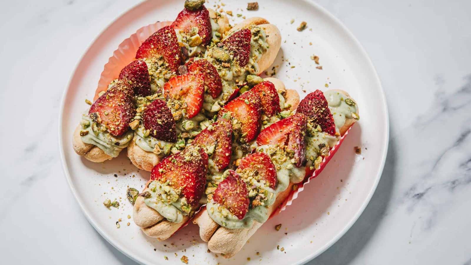 Three ladyfinger desserts topped with green cream, sliced strawberries, and chopped pistachios on a white plate.