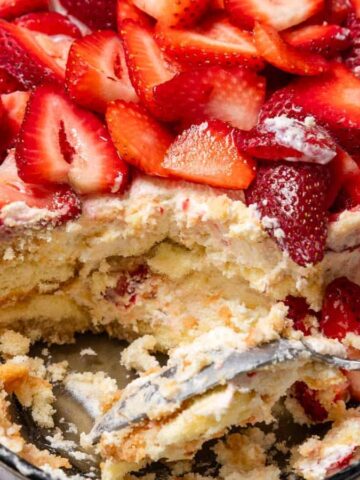 A partially eaten strawberry tiramisu with cream layers, sliced strawberries, and a spoon in the dish.