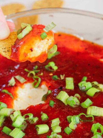 A hand dips a chip into a cheesy dip topped with red sauce and chopped green onions.