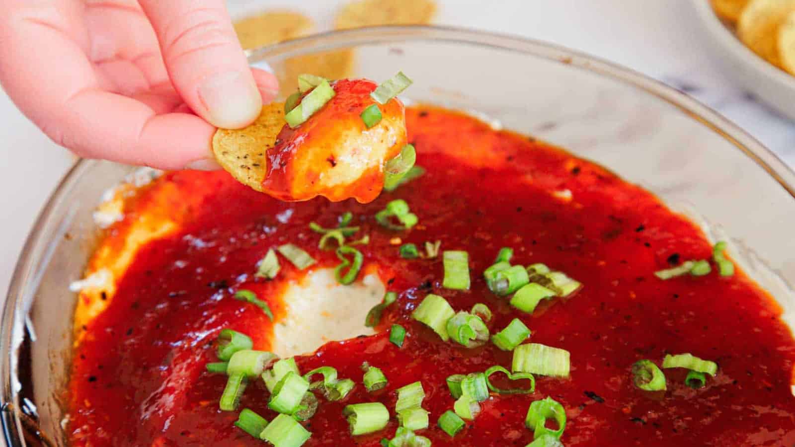 A hand dips a chip into a cheesy dip topped with red sauce and chopped green onions.