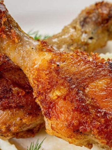 Two crispy fried chicken drumsticks on creamy mashed potatoes with a gold fork beside them.