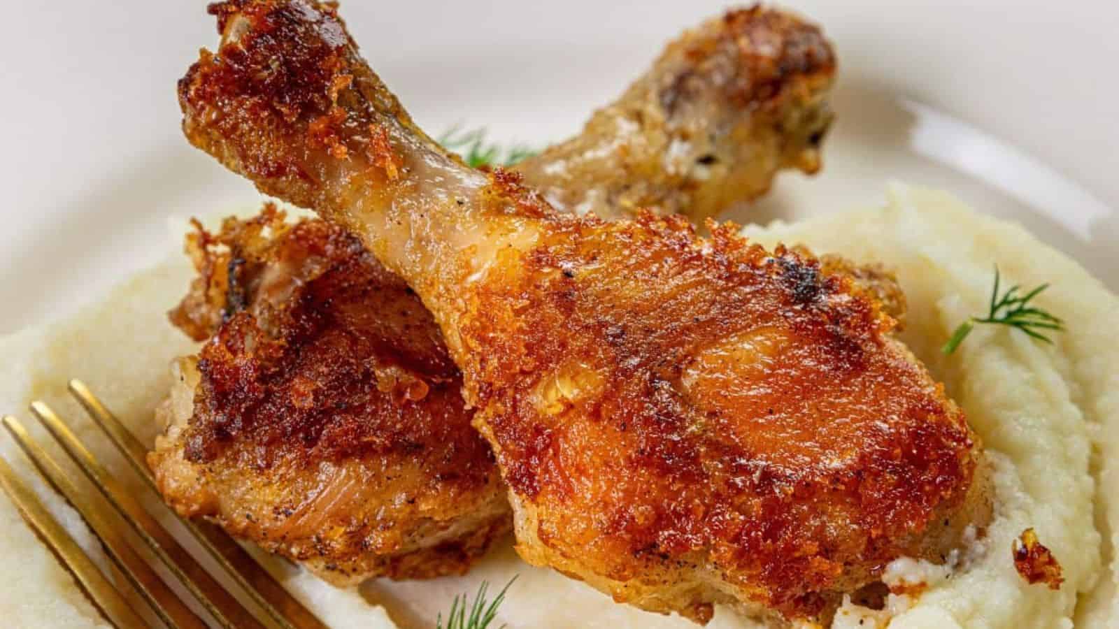 Two crispy fried chicken drumsticks on a bed of creamy mashed potatoes, garnished with dill. A fork is placed beside the dish. The chicken is golden brown and the potatoes have a smooth texture.