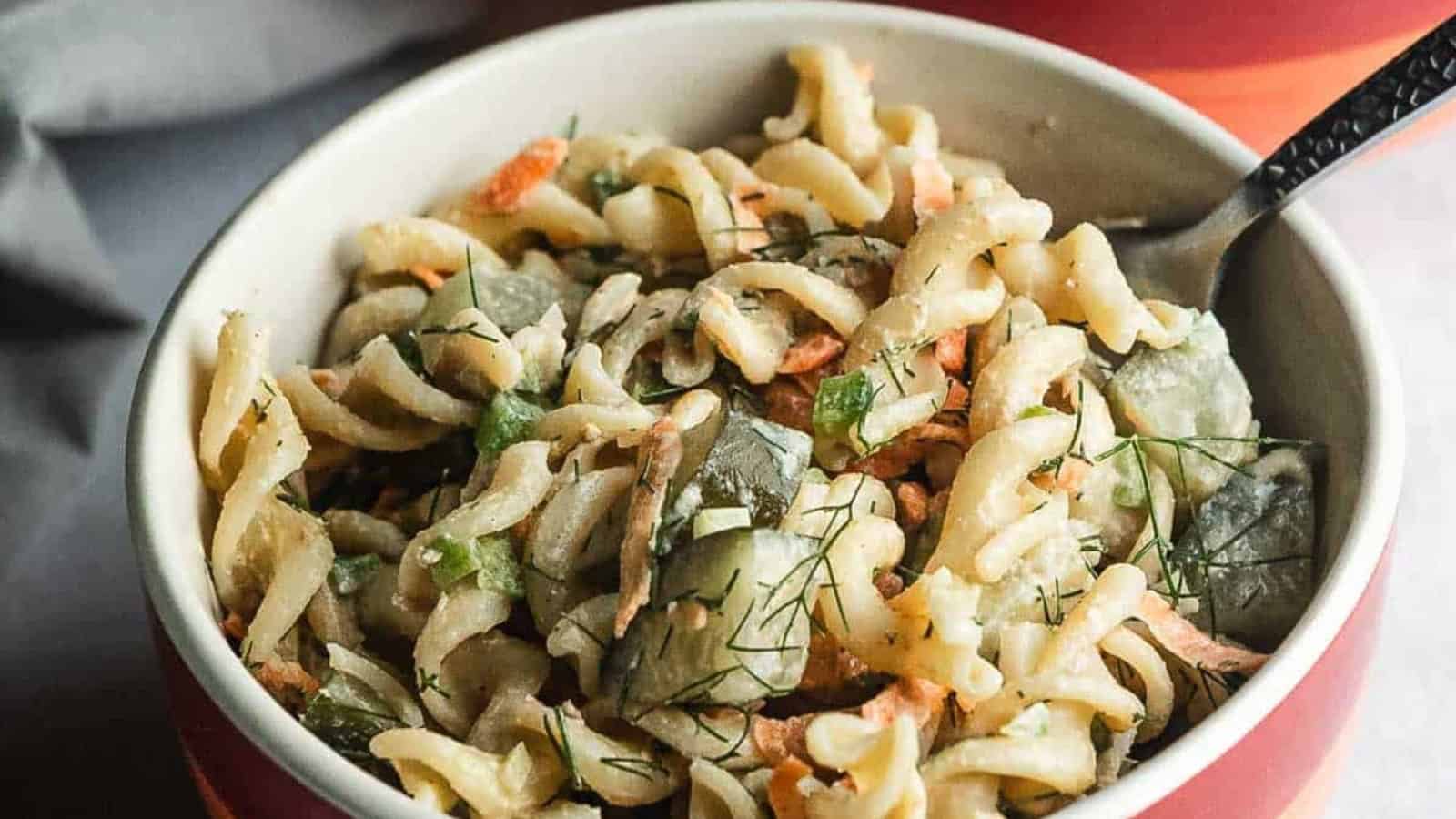 A bowl of rotini pasta salad with cucumbers, carrots, and herbs, with a fork inside.