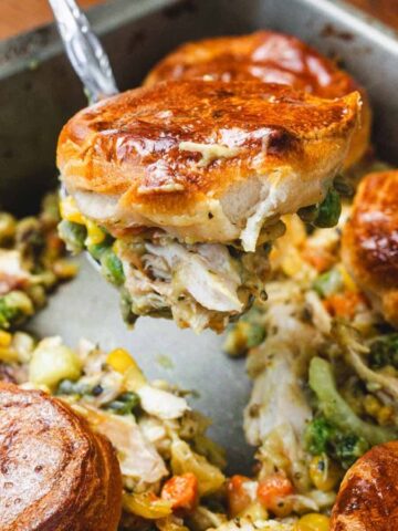 Golden biscuits on creamy chicken pot pie filling with vegetables in a baking pan, one biscuit being served.