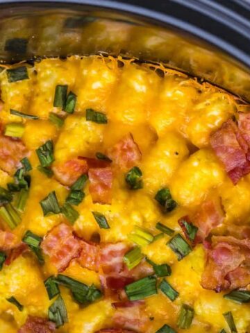 Cheesy tater tots topped with bacon bits and chopped chives in a slow cooker.