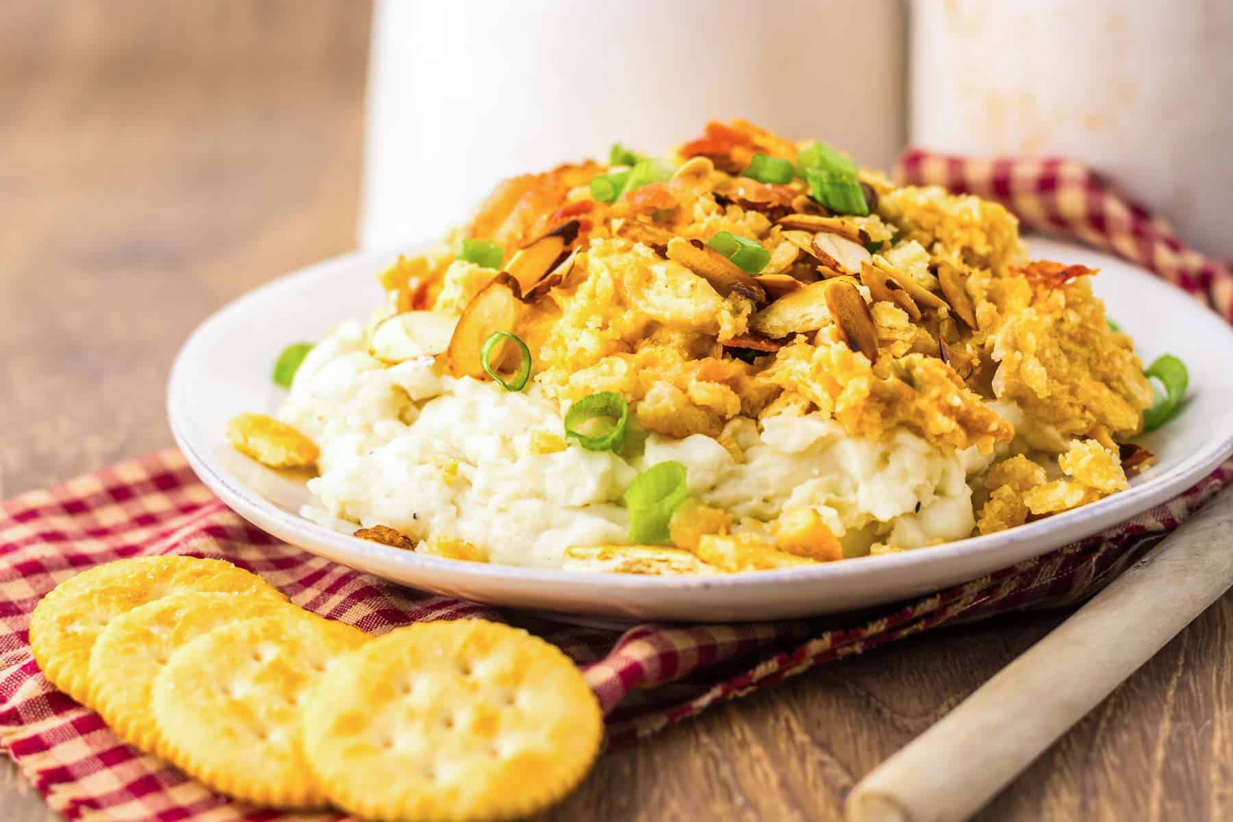 A plate of cheesy Neiman Marcus Chicken casserole with sliced almonds and green onions, served with crackers on a red plaid napkin.