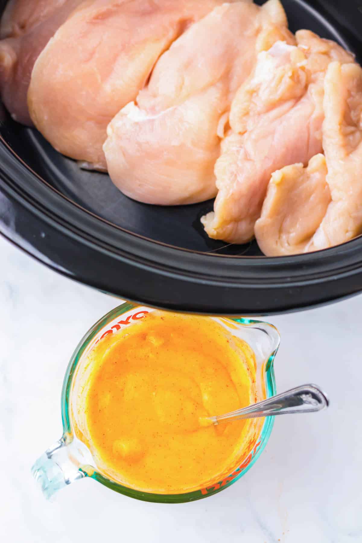 Raw chicken breasts in a slow cooker next to a glass measuring cup of yellow sauce with a spoon, perfect for making Neiman Marcus Chicken.