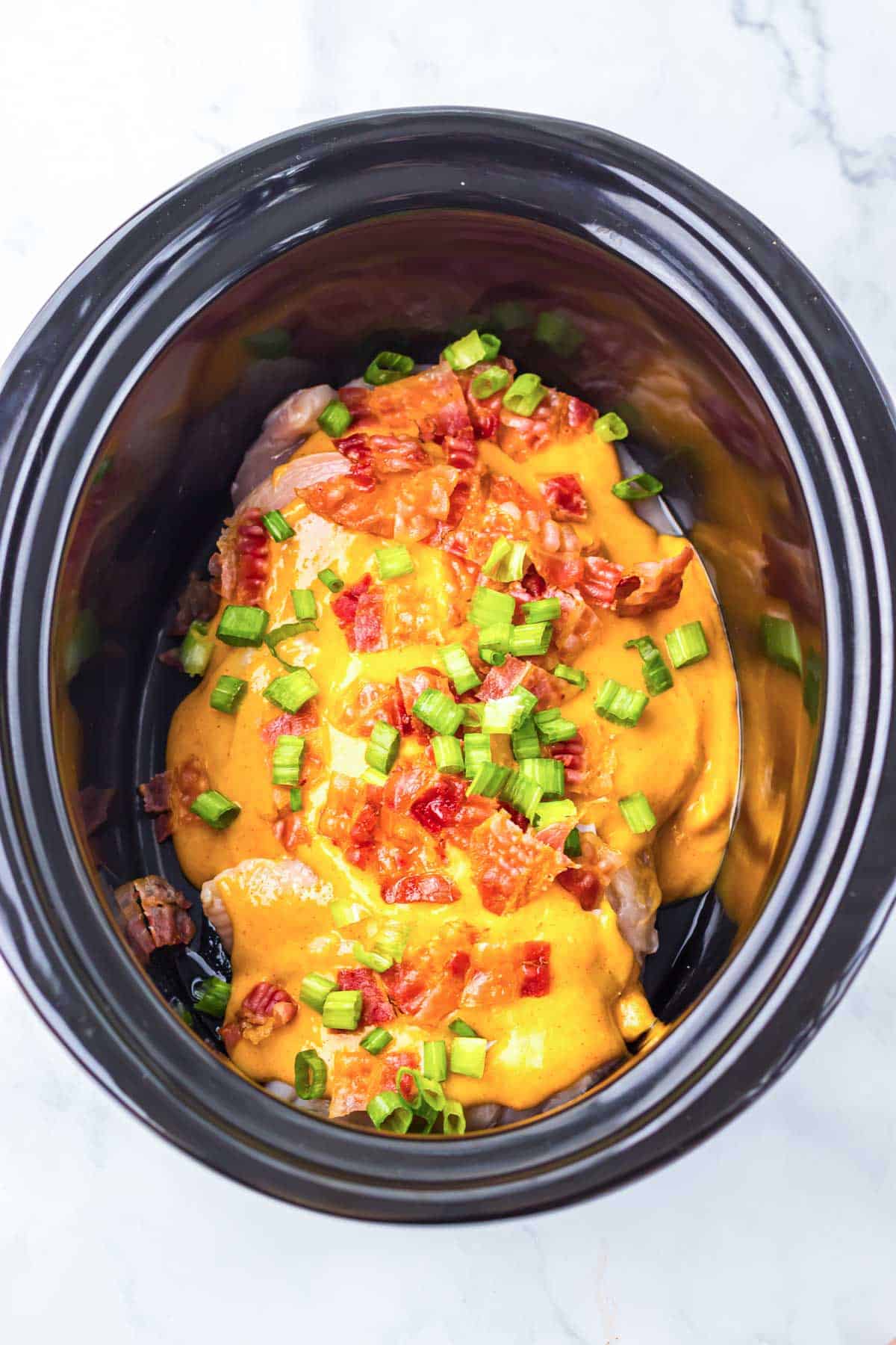 Neiman Marcus Chicken in a slow cooker, topped with orange sauce, chopped bacon, and green onions, sits ready on a marble surface.