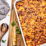 Golden baked macaroni and cheese in a white dish on a wooden board, with parsley and a wooden spoon nearby.