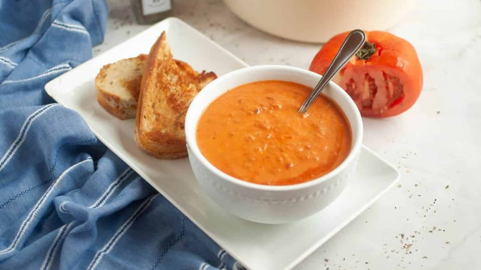 A bowl of tomato soup with a spoon, served with two slices of toasted bread on a white plate; a halved tomato and a blue cloth are nearby.