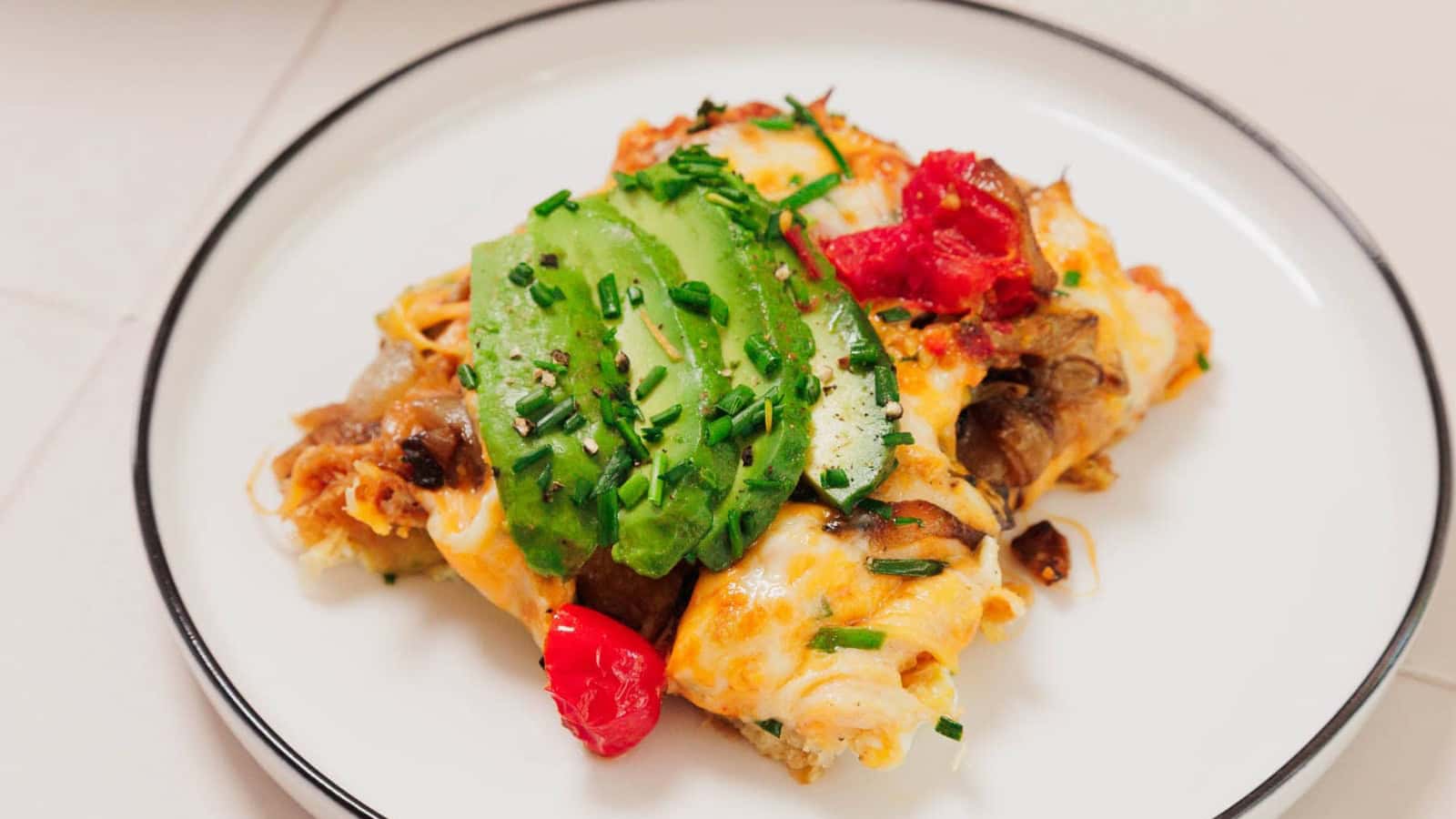Baked egg casserole with cherry tomatoes, avocado slices, and fresh herbs in a blue cast iron dish.