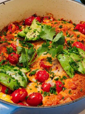 Baked casserole with avocado slices, cherry tomatoes, herbs, and cilantro in a blue dish.