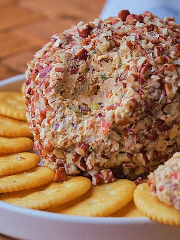 A round cheese ball coated in chopped nuts is placed on a white plate surrounded by round crackers, with a small portion of the cheese ball spread onto one cracker. A bowl of chopped nuts is nearby.