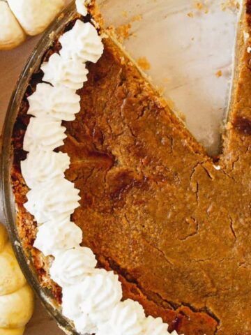Pumpkin pie with whipped cream and a missing slice, surrounded by orange and white pumpkins.