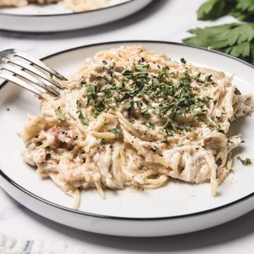 A plate of creamy chicken spaghetti, cooked in a slow cooker and garnished with chopped herbs, with a fork resting on the side.