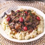 Shredded beef with gravy and cranberries served over mashed potatoes on a white plate with a fork.