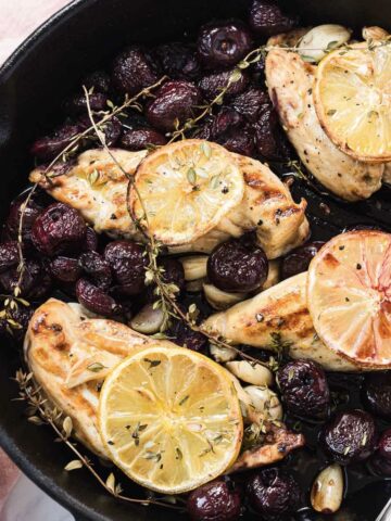 Grilled Cast Iron Chicken breasts with lemon slices, herbs, garlic, and roasted grapes in a black skillet.