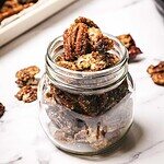 Glass jar filled with slow cooker candied pecans on a white surface, with more sugared pecans scattered nearby.