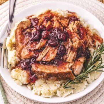 Tender Crockpot shredded pork loin with cranberry sauce served over creamy mashed potatoes, garnished with rosemary on a white plate.
