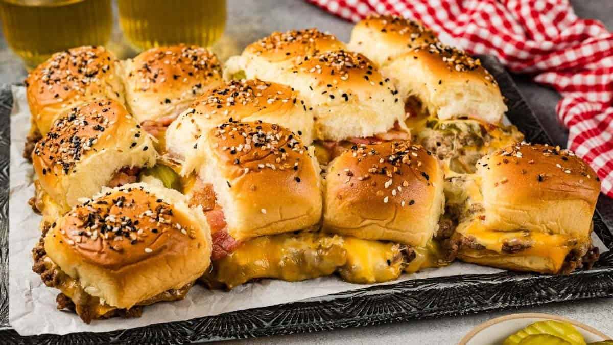 A tray of bacon cheeseburger sliders topped with sesame seeds and served on a dark surface, with a red checkered cloth and pickles nearby.
