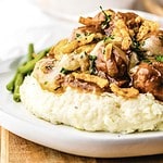 Mashed potatoes topped with slow cooker French onion meatballs, savory gravy, crispy onions, and herbs, served with green beans on a white plate.