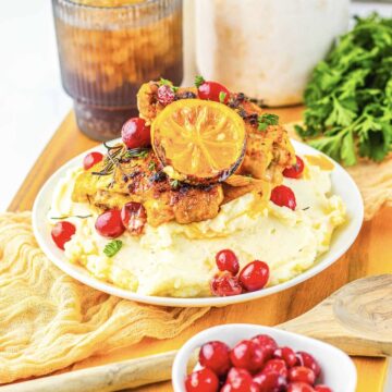 Roasted Cranberry Chicken with lemon sits atop mashed potatoes, garnished with herbs on a white plate.