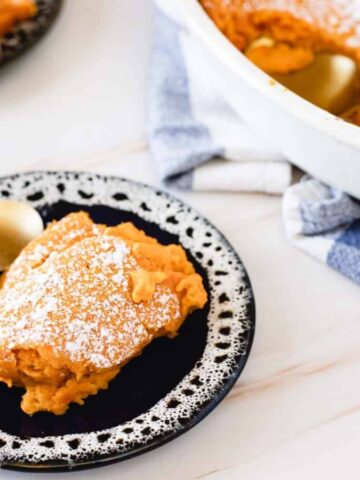 A slice of sweet potato casserole topped with powdered sugar on a black plate with a gold spoon.