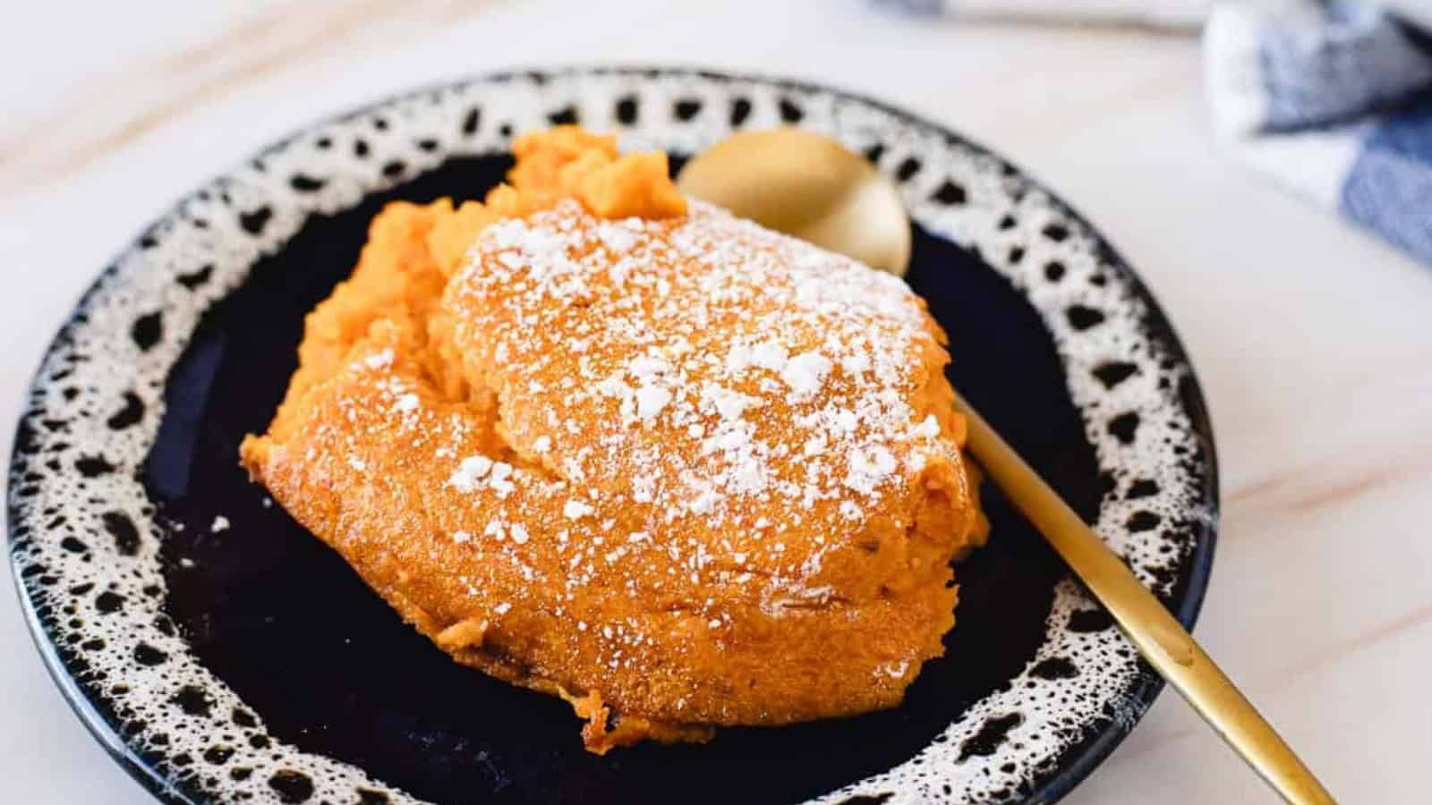 A slice of sweet potato souffl&eacute; topped with powdered sugar on a black and white plate with a gold spoon.