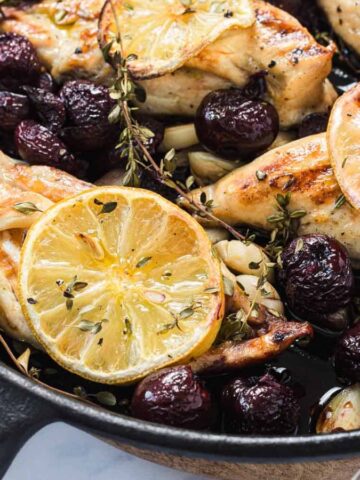 Skillet with roasted chicken breasts, lemon slices, grapes, garlic, and fresh herbs.