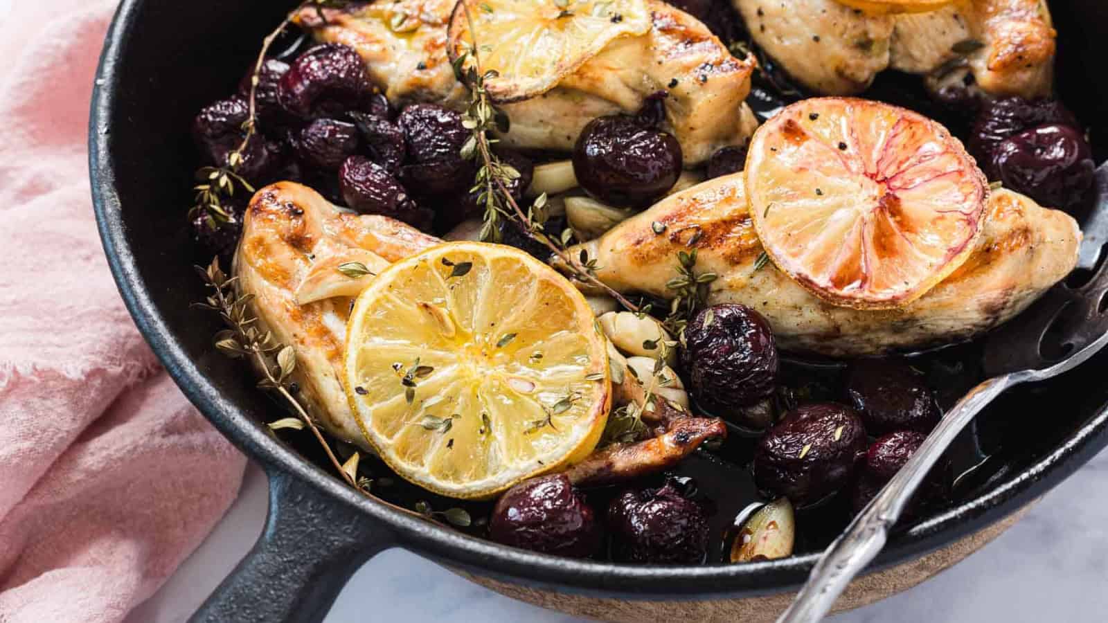 Skillet with roasted chicken breasts, lemon slices, grapes, garlic, and fresh herbs.