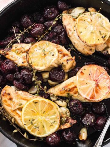Chicken breasts with lemon slices, roasted grapes, garlic, and herbs in a black skillet on a marble surface.