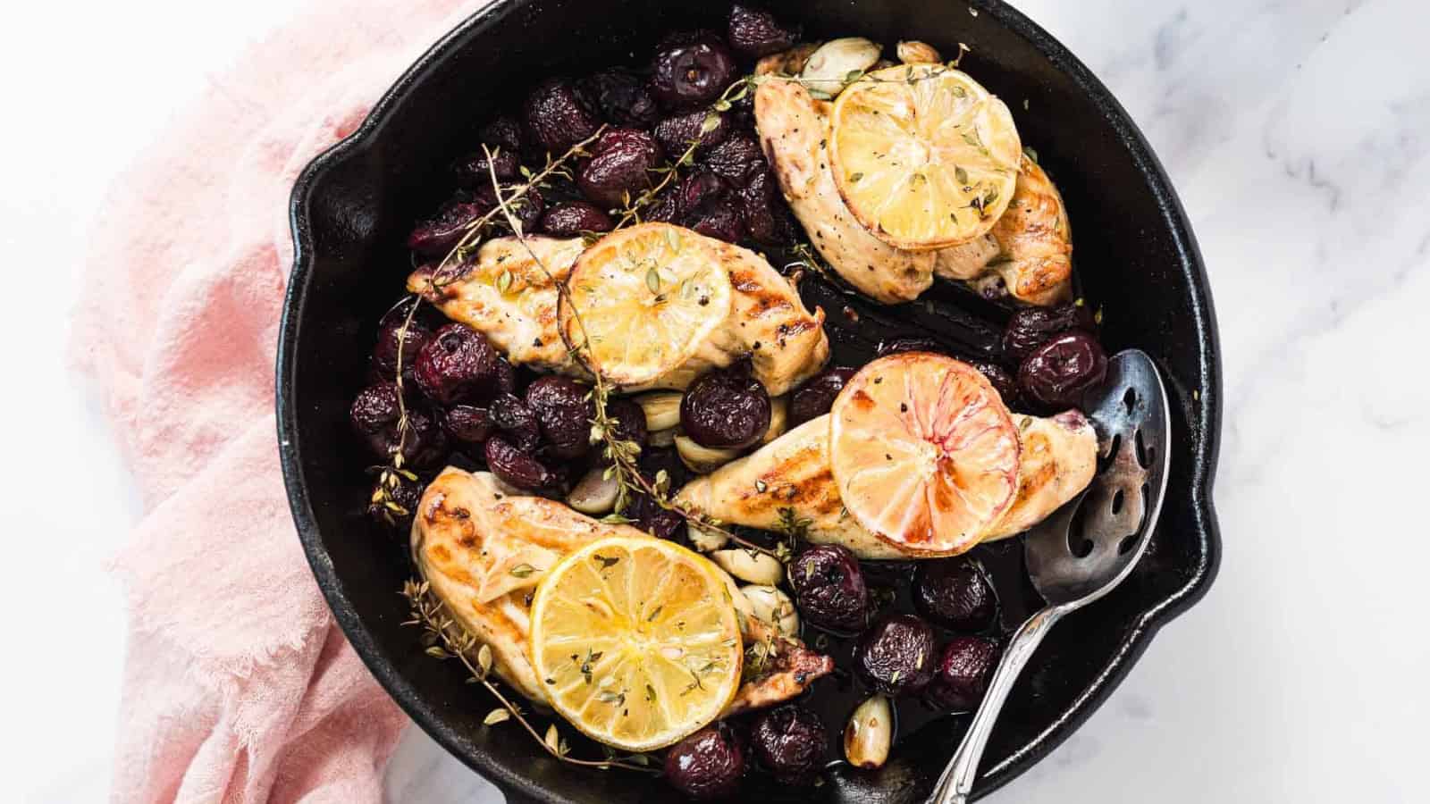 Skillet with roasted chicken breasts, lemon slices, grapes, herbs, and garlic cloves.