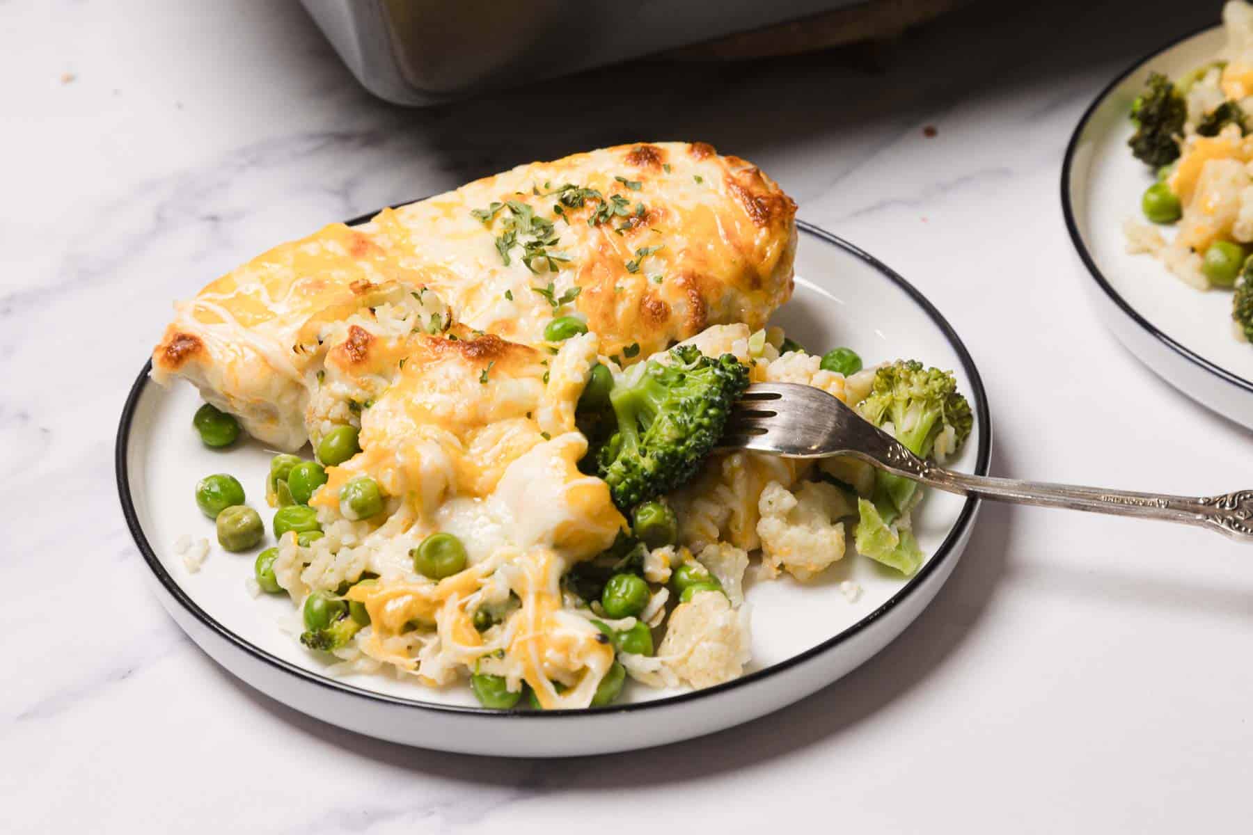 A plate of cheesy Chicken and Rice Casserole with green peas, cauliflower, and broccoli sits on a white table.