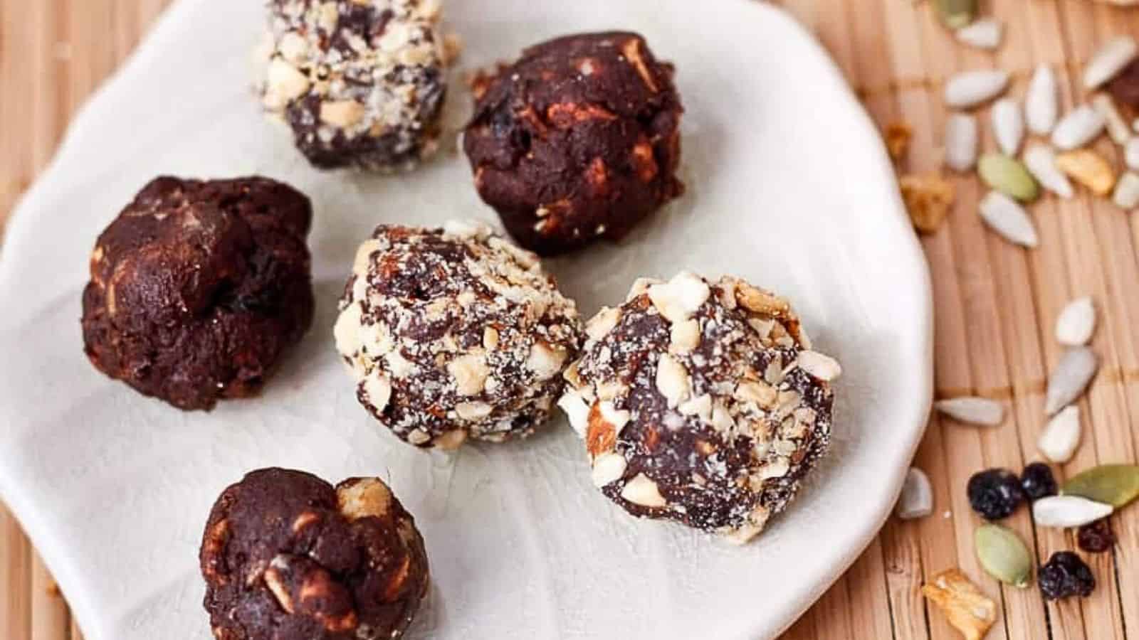 Five chocolate energy balls, some coated in chopped nuts, on a white plate with seeds scattered nearby.