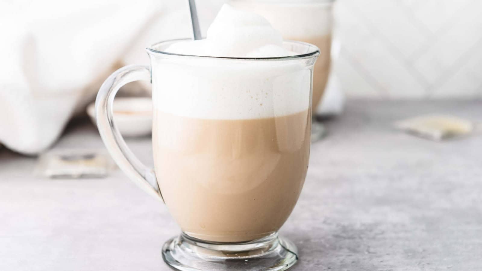 A glass mug of frothy chai latte on a light surface, with another mug blurred in the background.